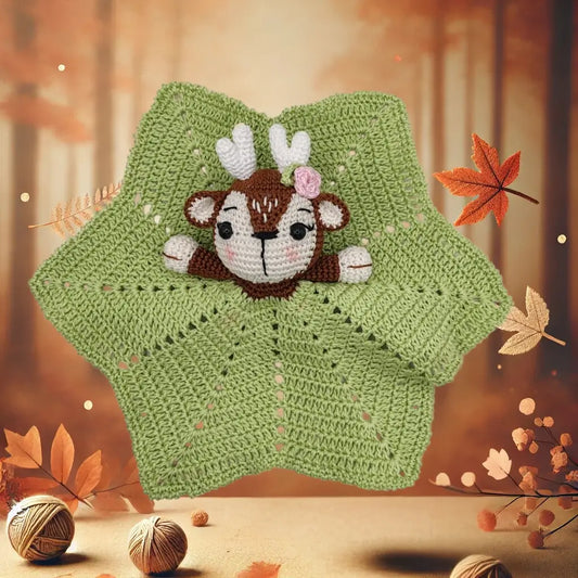 An amigurumi doudou featuring a fawn with white antlers, a decorative rose on its head, and rosy cheeks. The body of the doudou is a green, star-shaped blanket crocheted with lacy patterns. The background evokes an autumn atmosphere, with orange and brown leaves, yarn balls, and a blurred forest in the distance.