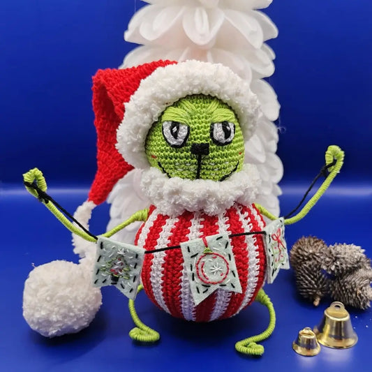 An amigurumi depicting a green character inspired by the Grinch, featuring a grumpy face with well-defined details. It wears a red Christmas hat with a fluffy white trim and a red-and-white striped body. Its hands hold a decorative garland. The background is blue, adorned with Christmas decorations like golden bells and a stylized white tree.